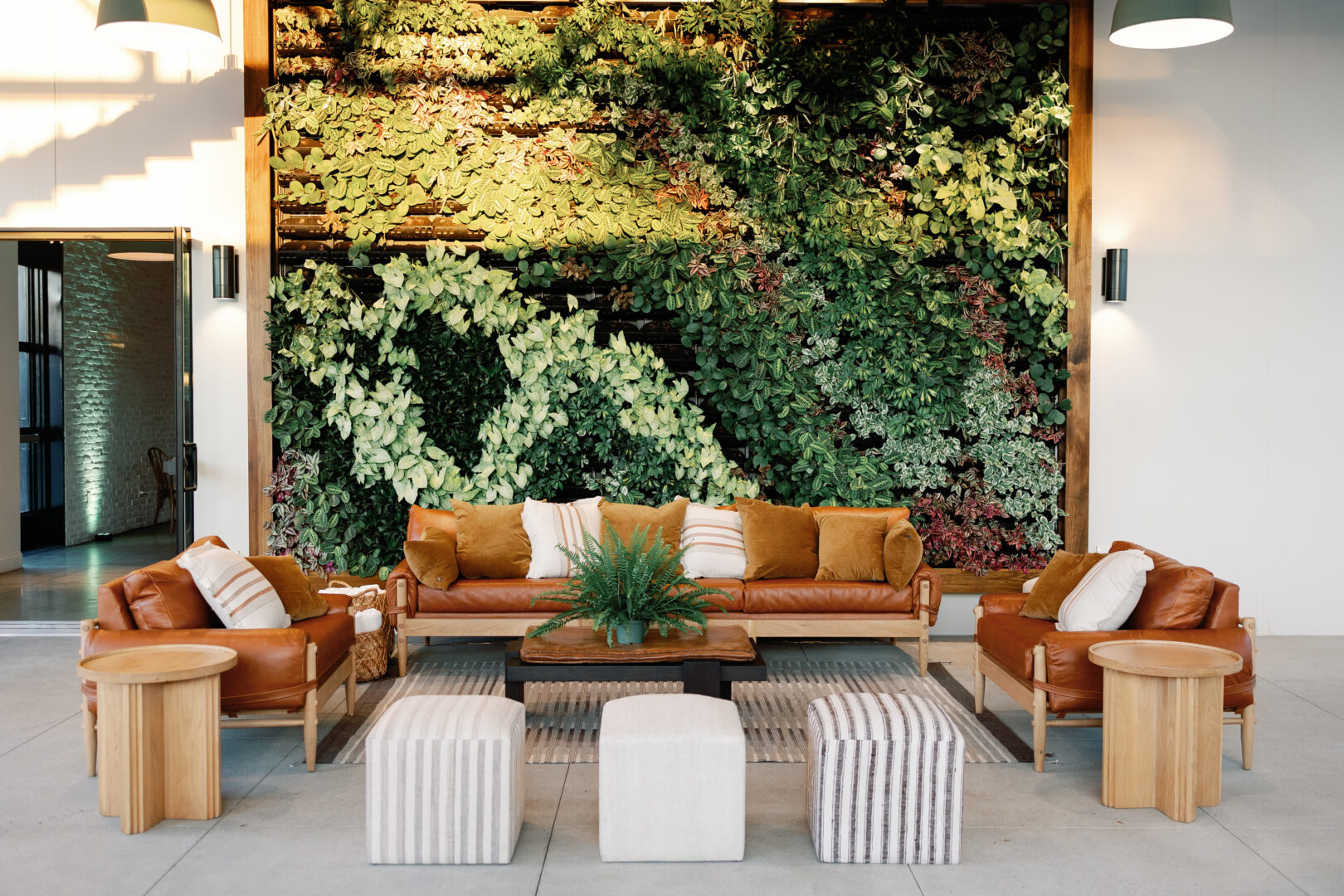 Modern lounge area with brown leather sofas, striped ottomans, wooden tables, and a coffee table, set in front of a large vertical green plant wall. Pillows and a potted plant complete the cozy, natural look.