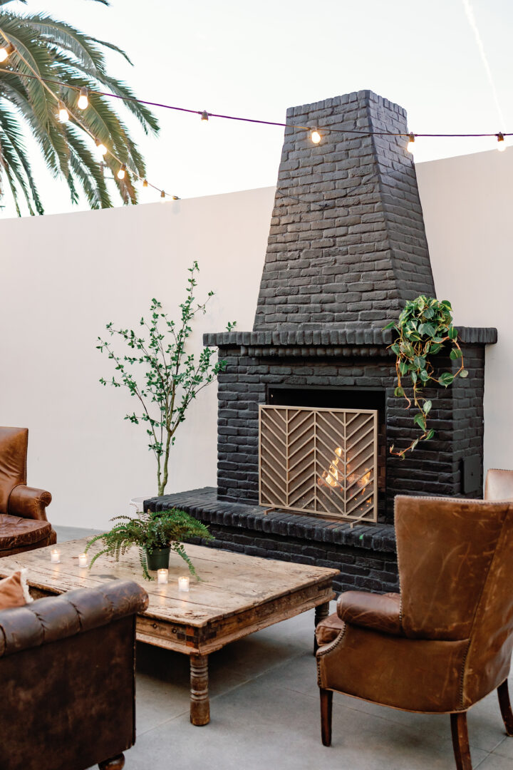 Outdoor patio with a black brick fireplace, wooden coffee table, leather chairs, potted plants, and string lights overhead. Palm tree and white wall in the background. Cozy and inviting atmosphere.