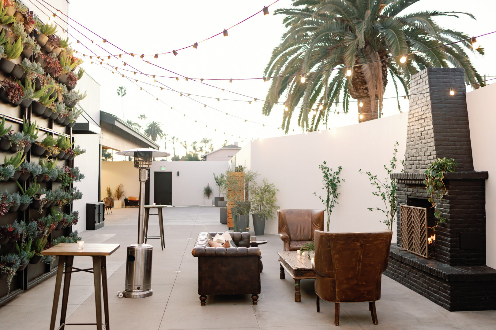 Outdoor patio with seating, leather chairs, a brick fireplace, string lights overhead, potted plants, and a vertical garden on a white wall. Palm trees are visible in the background.