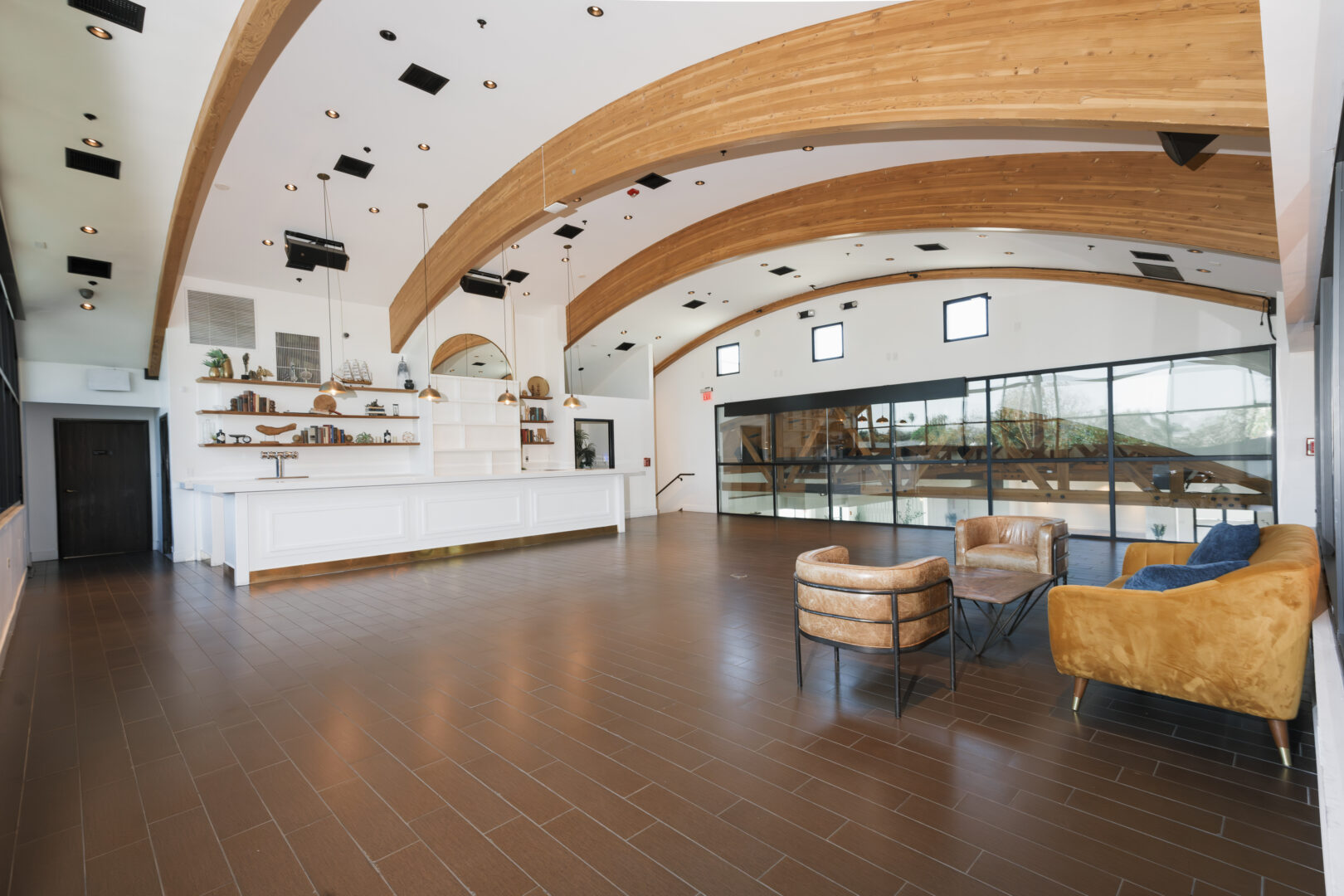 Spacious modern lobby with arched wooden beams, white walls, brown tile floor, a white reception desk, shelves with decor, and seating area with leather chairs and a tan sofa. Large windows let in natural light.