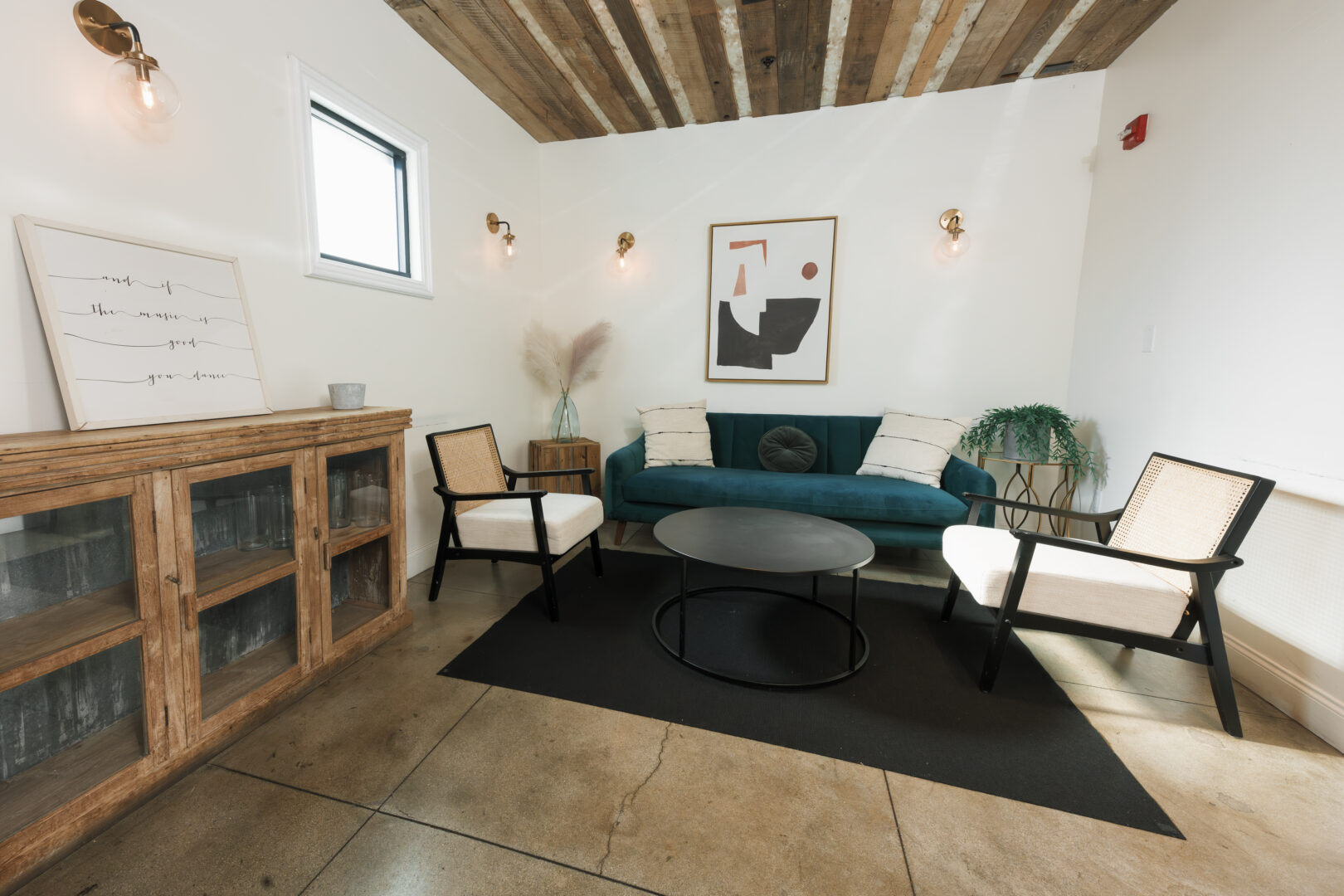 Modern living room with a green sofa, two chairs, a round coffee table, abstract wall art, and a wooden cabinet. The room has white walls, exposed wood ceiling, and a small window.