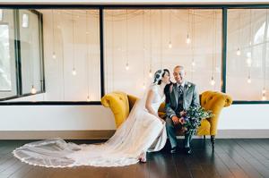 A bride and groom smile and sit next to each other on a single mustard couch